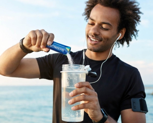 man pouring electrolyte stick pack powder into a shaker bottle while out running near the water